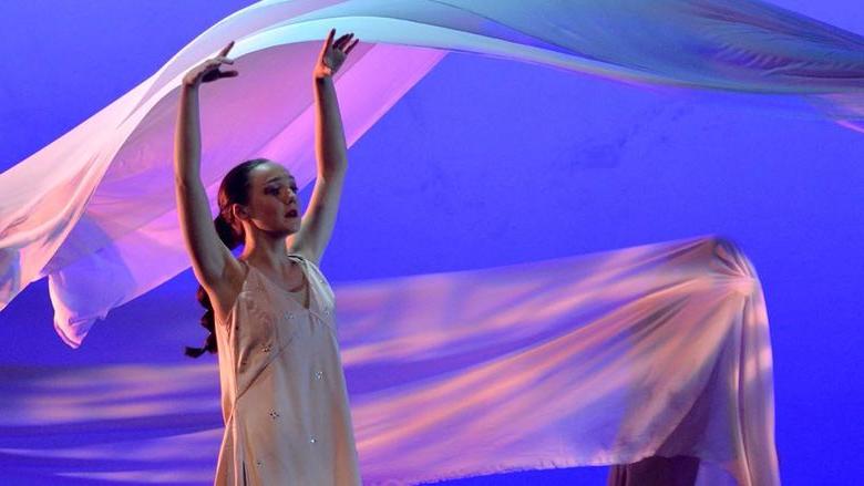 A dancer in the foreground with her arms raised in the air while a piece of fabric flows in the air behind her